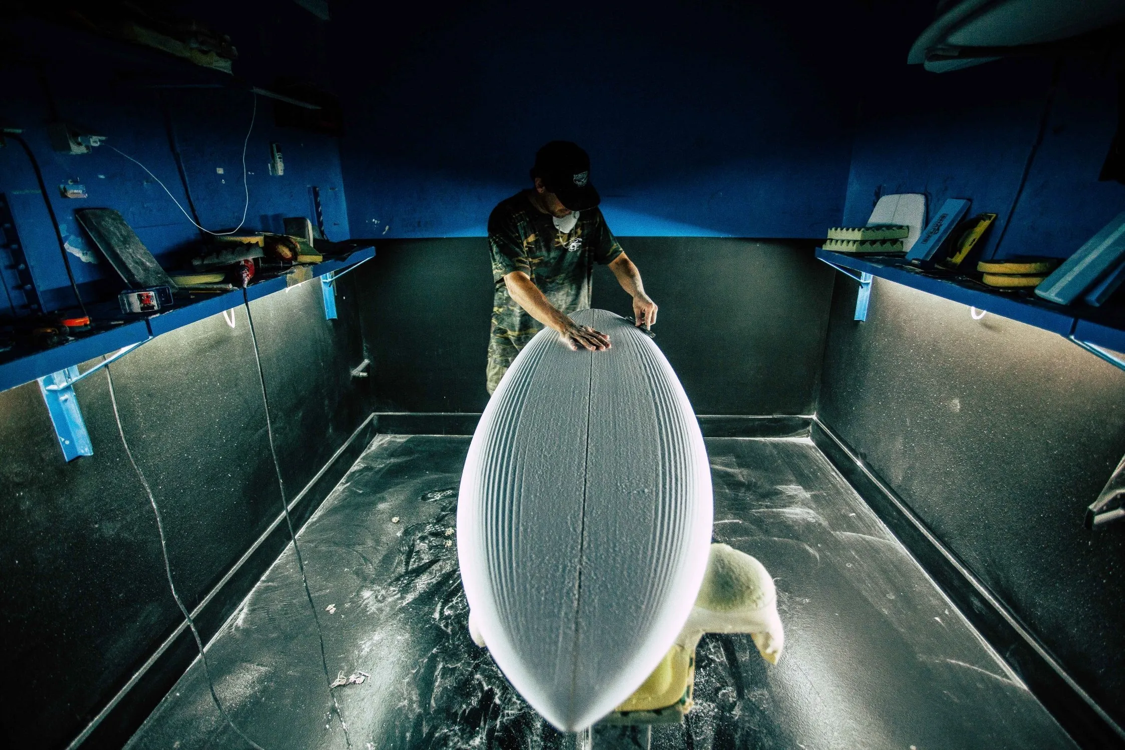 Man working on surfboard in workshop, sanding and shaping.